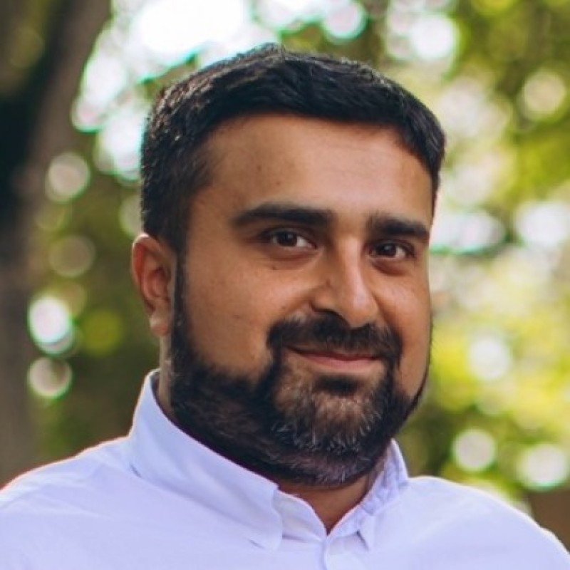 Ein Mann mit kurzen dunklen Haaren und Bart, der ein weißes Hemd mit Kragen trägt, lächelt sanft, während er im Freien steht, während die grünen Bäume im Hintergrund verschwimmen, und verkörpert so das sympathische Vertrauen eines echten Kopiloten.