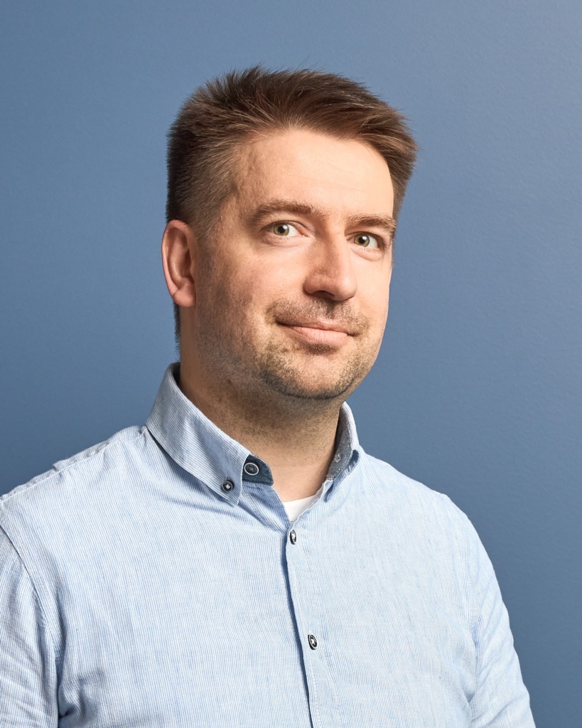 Mężczyzna z krótkimi brązowymi włosami i jasnym zarostem ma na sobie jasnoniebieską koszulę zapinaną na guziki i stoi na prostym niebieskim tle, patrząc lekko w bok z neutralnym wyrazem twarzy.