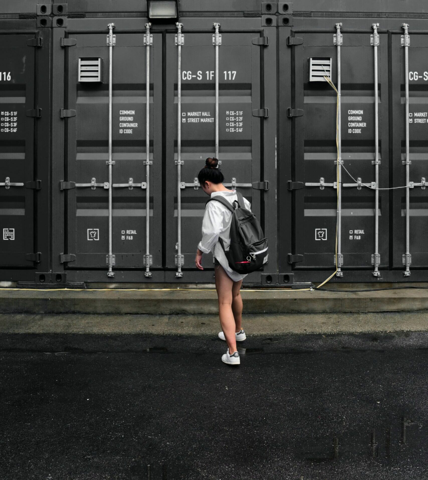 Osoba z czarnym plecakiem i białymi trampkami idzie po mokrym chodniku obok dużych, szarych kontenerów transportowych z etykietami i metalowymi zamkami.