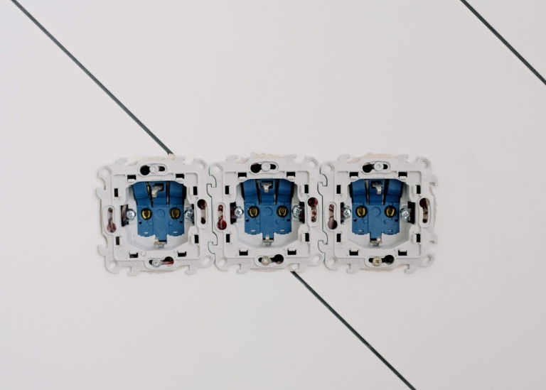 Trzy puste białe skrzynki elektryczne z niebieskimi wnętrzami są zainstalowane obok siebie na białej ścianie z cienkimi ukośnymi liniami.