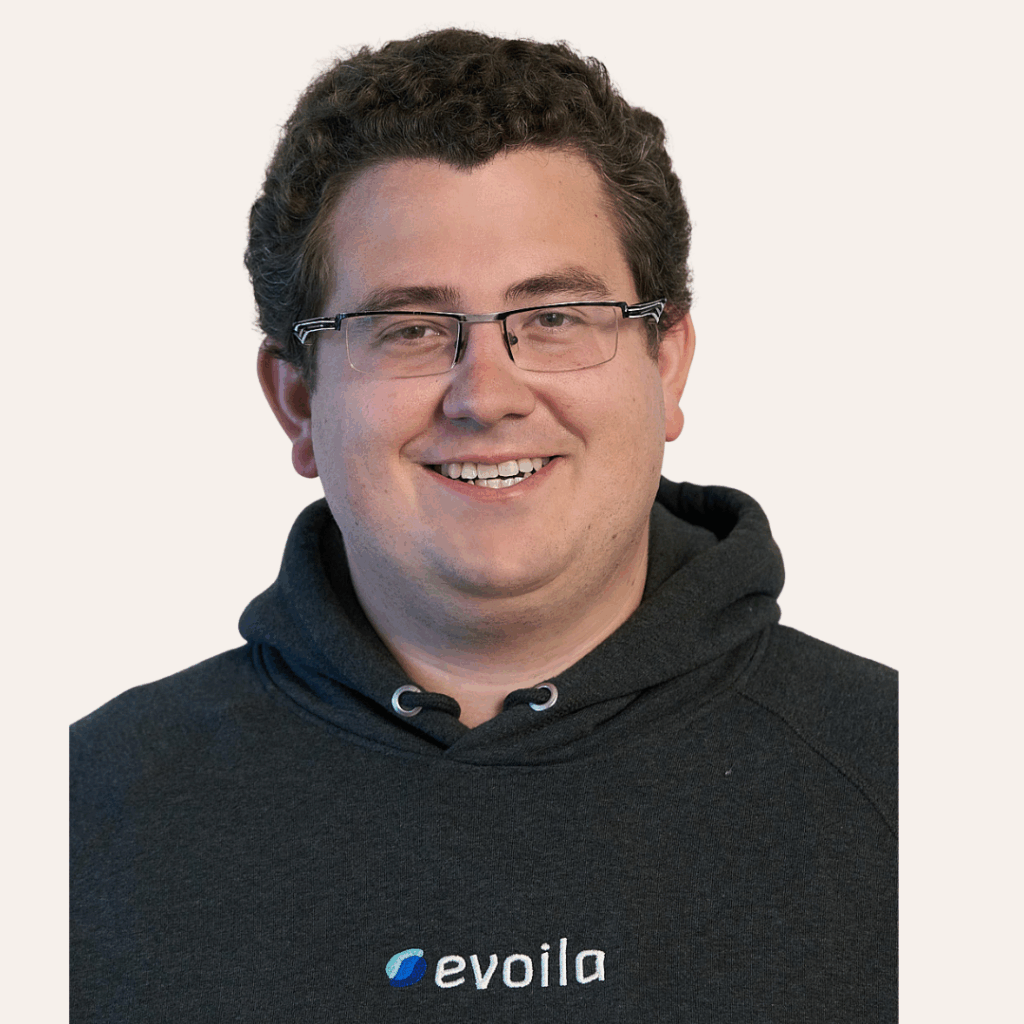 Ein Mann mit kurzen lockigen braunen Haaren, Brille und einem schwarzen Kapuzenpulli mit dem evoila"-Logo lächelt vor einem schlichten hellen Hintergrund in die Kamera, bereit, evoila auf der Microsoft Ignite 2025 zu vertreten.