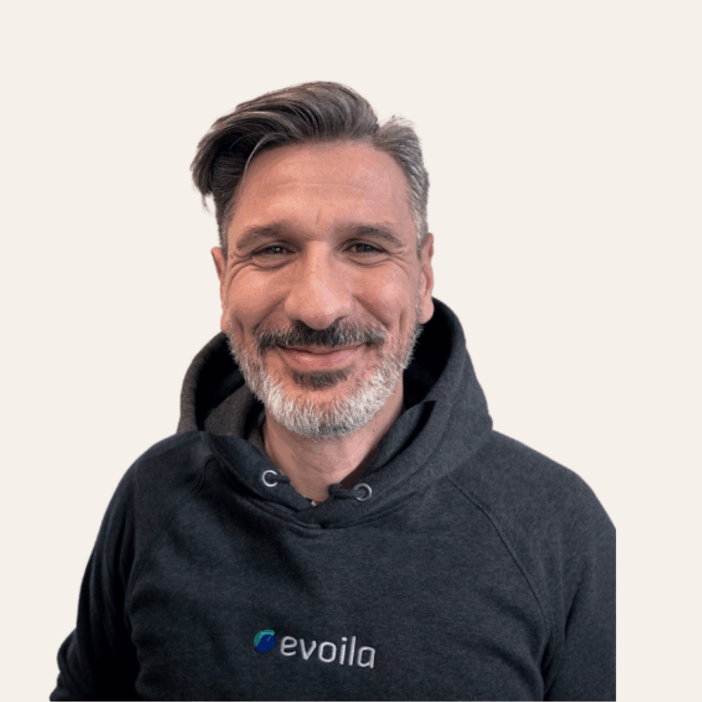 Ein Mann mit kurzen grauen Haaren und einem Bart lächelt in die Kamera. Er trägt einen dunklen Kapuzenpulli mit dem evoila-Logo darauf und steht vor einem schlichten hellen Hintergrund, bereit für die Microsoft Ignite 2025.
